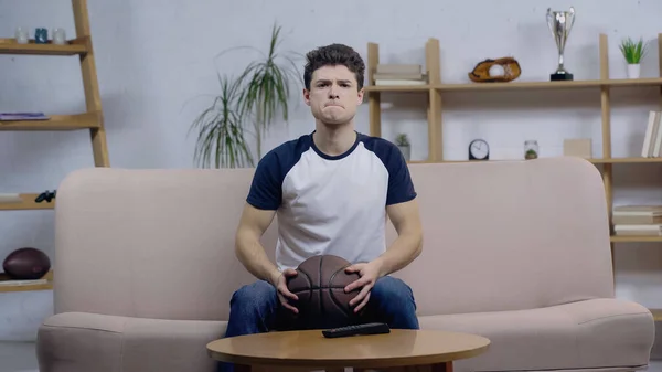 Worried basketball fan holding ball while watching match on tv at home — Stockfoto
