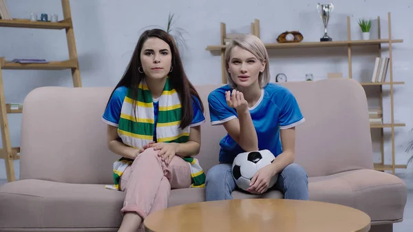 Беспокойная брюнетка и блондинка, смотрящие футбольный чемпионат на диване дома — стоковое фото