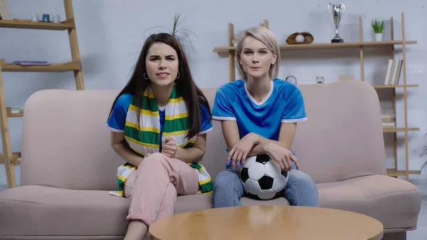 Tense and concentrated women sitting on sofa with soccer ball and watching game on tv — Stock Photo