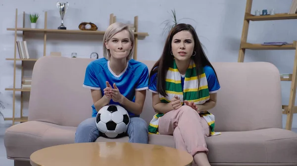 Worried sport fans with soccer ball and striped scarf watching football match on home tv — Stock Photo