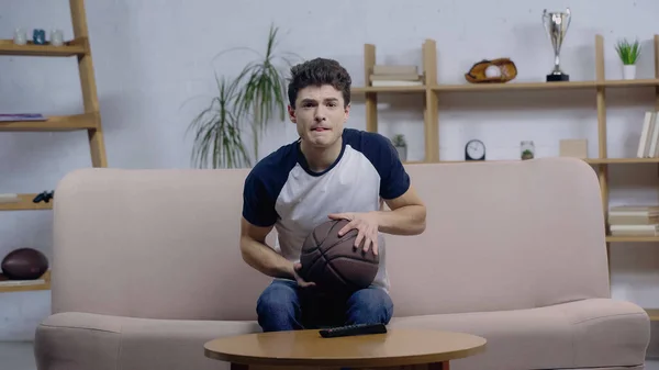 Напружений фанат спорту сидить на дивані з м'ячем під час перегляду баскетбольної гри на домашньому телебаченні — стокове фото