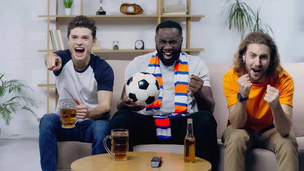 Aufgeregter Mann zeigt mit dem Finger in die Nähe multiethnischer Freunde und zeigt beim Zuschauen eines Fußballspiels Siegergeste — Stockfoto
