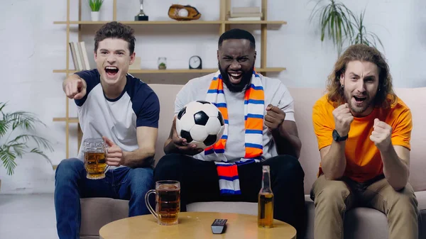 Hombre excitado señalando con el dedo cerca de amigos multiétnicos mostrando gesto de victoria mientras mira el partido de fútbol - foto de stock