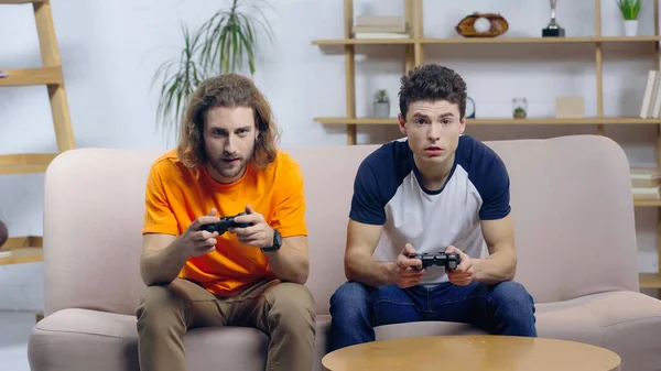 KYIV, UKRAINE - DECEMBER 22, 2021: young concentrated friends playing video game with joysticks at home — Foto stock