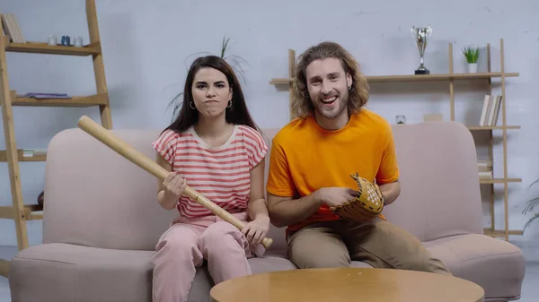 Angry woman holding baseball bat near happy man while watching match on tv at home — Fotografia de Stock