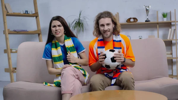 Smiling man holding soccer ball while watching match on tv near nervous woman — Stockfoto