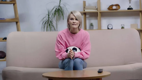 Donna tesa seduta sul divano con pallone da calcio e guardando il campionato in tv — Foto stock