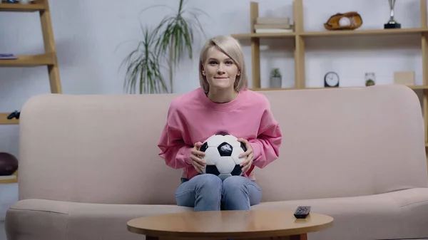 Smiling football fan woman holding ball while watching championship on sofa at home — Stockfoto