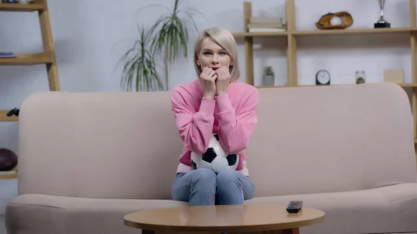 Worried sport fan woman with soccer ball watching football match on couch at home — Foto stock