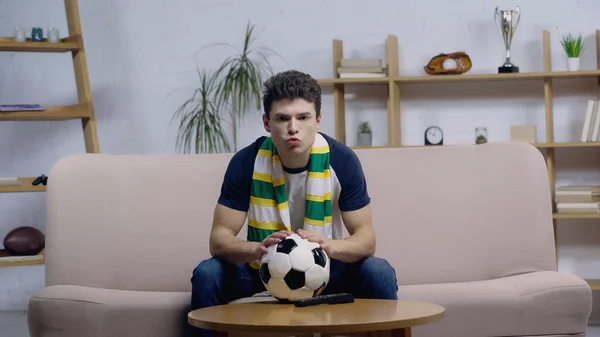 Emocionado joven fanático del fútbol sentado en el sofá cerca de la pelota de fútbol en la mesa de café y viendo el juego en la televisión - foto de stock