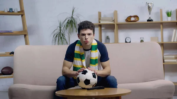 Thrilled young football fan sitting on couch near soccer ball on coffee table and watching game on tv — Foto stock