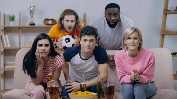 Anxious multicultural sport fans watching football game near beer and chips at home — Stock Photo