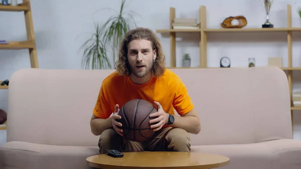 Fan de sport concentré tenant le ballon tout en regardant le match de basket sur la télévision à domicile — Photo de stock