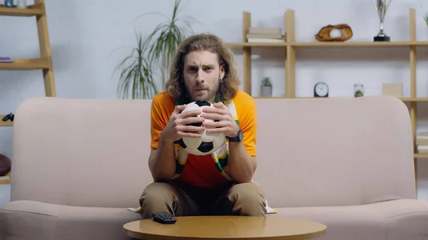 Tense man holding soccer ball while watching football match on tv at home — Stockfoto