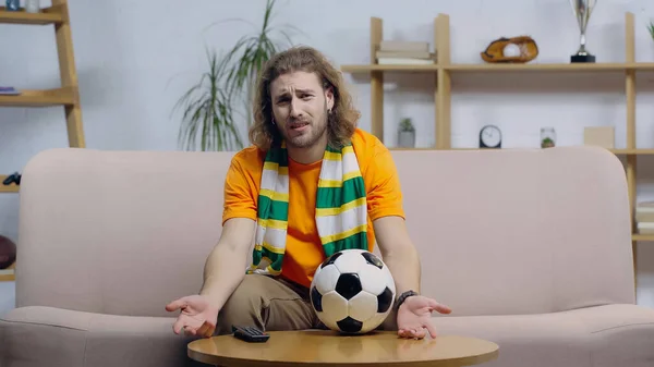 Frustrated football fan gesturing while watching match near ball on coffee table — Stock Photo