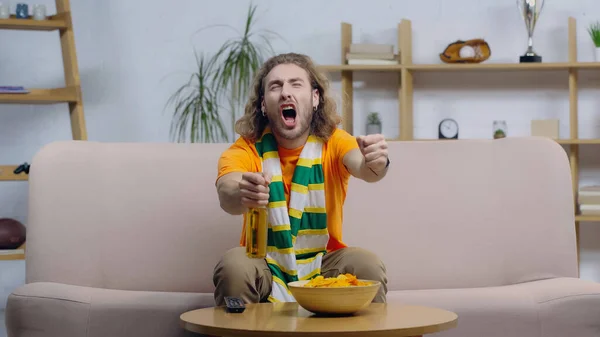 Excited sport fan screaming and showing win gesture while sitting on couch ner beer and chips — Stock Photo