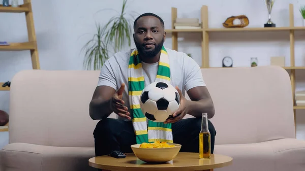 African american man in striped scarf holding soccer ball while watching championship near chips and beer — Stock Photo