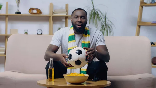 Excited african american man holding soccer ball while watching football championship near beer and chips — Stock Photo