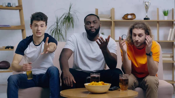 Dissatisfied sport fans watching championship and pointing with hands while sitting on couch — Stock Photo