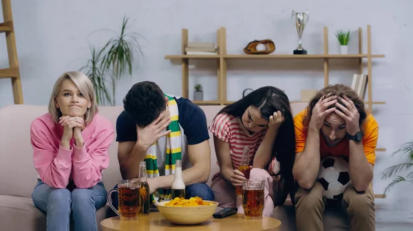 Frustrated football fans sitting with bowed heads near beer and chips at home — Stock Photo