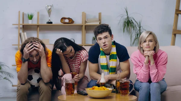 Frustrated friends sitting near coffee table with beer and chips while watching football match on tv — Stock Photo