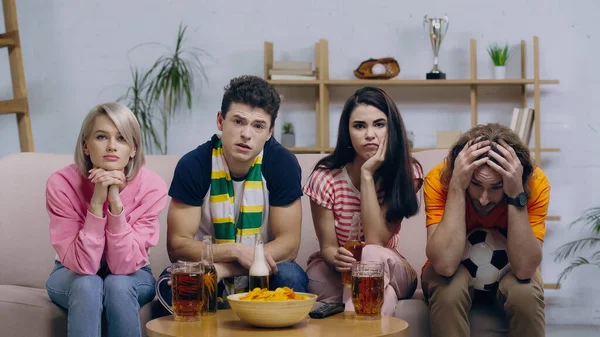 Unhappy football fans watching match on tv while sitting on couch near beer and chips — Stock Photo
