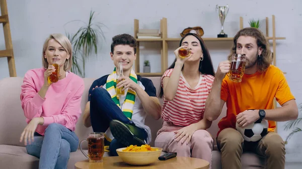 Fãs de futebol feliz beber cerveja enquanto assiste campeonato no sofá em casa — Fotografia de Stock