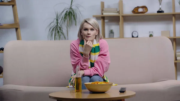 Skeptical sport fan in striped scarf watching match on tv near beer and chips — Stock Photo