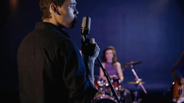 Cantante joven cantando en el micrófono cerca de banda de música borrosa - foto de stock