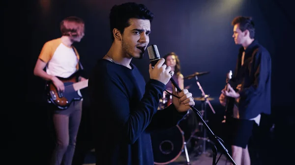 Joven cantante interpretando canción con banda de música borrosa en el escenario - foto de stock