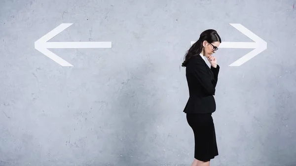 Pensive businesswoman in glasses thinking near arrows on grey — Photo de stock