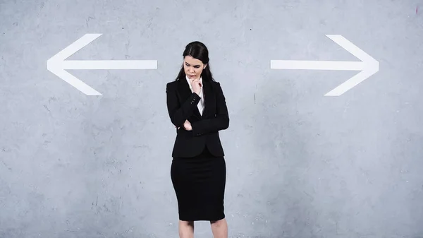 Pensive businesswoman in suit standing near arrows on grey — Photo de stock