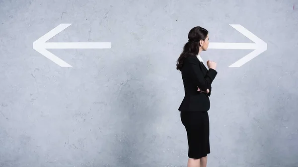 Thoughtful businesswoman in suit making choice while looking at arrows on grey — Stock Photo