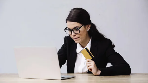 Бізнес-леді в окулярах тримає кредитну картку біля ноутбука ізольовано на сірому — стокове фото