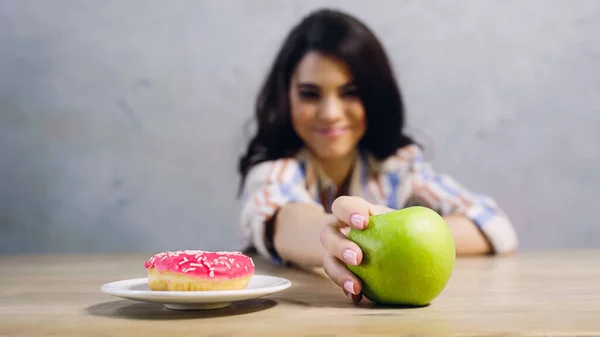 Mujer borrosa tomando manzana verde cerca de donut en gris - foto de stock
