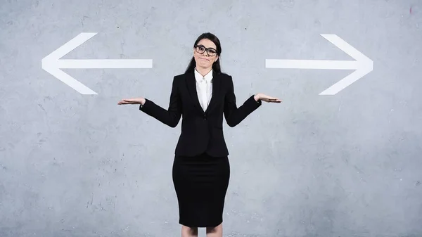 Confused businesswoman in glasses showing shrug gesture near arrows on grey — Stock Photo