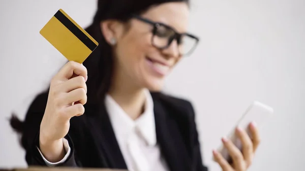 Happy and blurred businesswoman doing online shopping while holding smartphone and credit card isolated on white — Foto stock