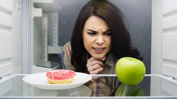 Brunette woman choosing between apple and glazed doughnut in fridge — Stockfoto