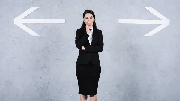 Pensive brunette businesswoman in suit standing near arrows on gray - foto de stock
