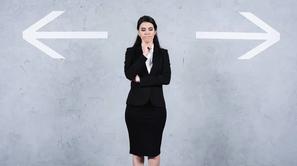 Pensativo morena mujer de negocios en traje de pie cerca de flechas en gris - foto de stock