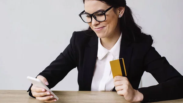 Mujer de negocios complacida sosteniendo teléfono inteligente y tarjeta de crédito aislada en blanco - foto de stock