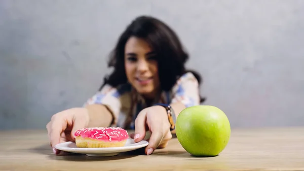 Feliz mujer tomando plato con donut dulce cerca de manzana en la mesa en gris - foto de stock