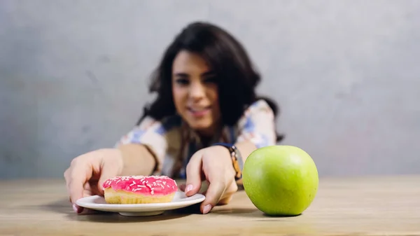 Happy woman taking plate with sweet doughnut near apple on table on grey — Stockfoto