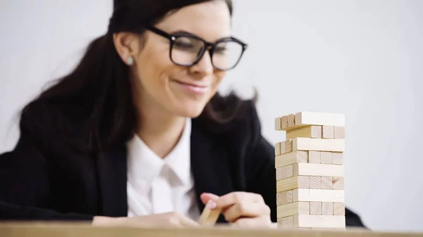 Blurred businesswoman in glasses smiling while playing blocks wood tower game isolated on grey — стоковое фото