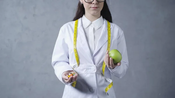 Частковий вигляд брюнетки дієтолога в білому пальто порівнюючи яблуко і вітаміни на сірому — стокове фото