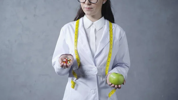 Partial view of nutritionist in white coat comparing apple and vitamins on grey — Stockfoto