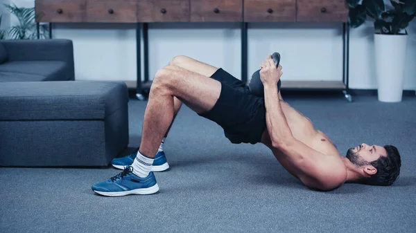 Side view of muscular sportsman training with kettlebell on floor at home — стоковое фото