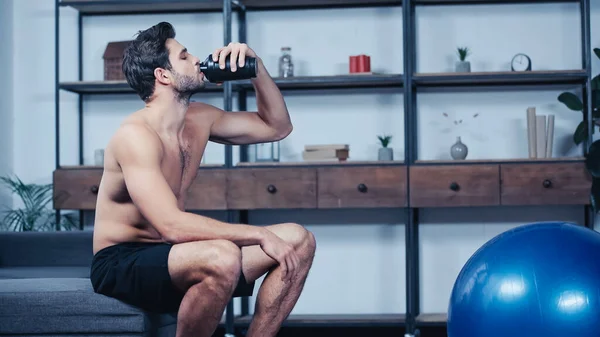 Hemdloser Sportler sitzt auf Sofa und trinkt Wasser aus Sportflasche — Stockfoto