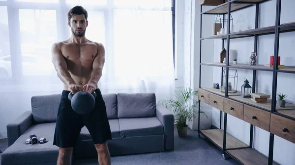 Shirtless sportsman training with kettlebell near sofa with dumbbells at home — Stock Photo