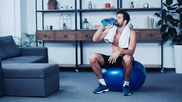 Tired sportsman with towel sitting on fitness ball and drinking water from sports bottle — Stock Photo